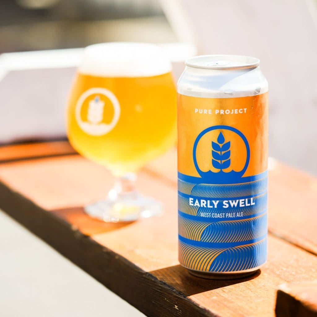 Can of Pure Project Early Swell West Coast pale ale in the foreground with a glass of the beer bathed in sunshine in the background