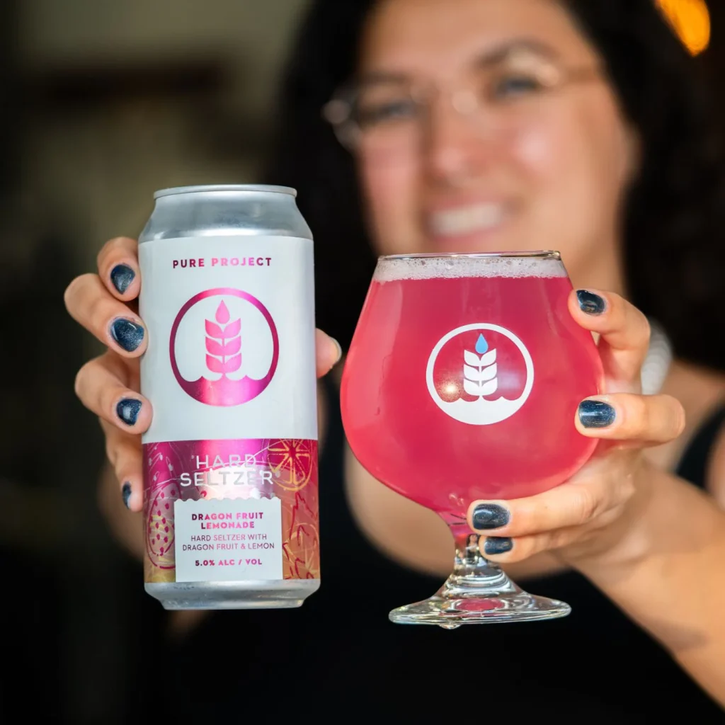 Woman holding a can of Pure Project Dragon Fruit Lemonade Hard Seltzer and a glass filled with a pink hard seltzer